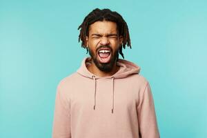 Stressed young good looking dark skinned brunette guy with dreadlocks frowning his face while screaming with wide mouth opened, posing over blue background with closed eyes photo