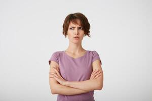 Portrait of frowning short-haired lady in blank t-shirt, trying to remember what offended her boyfriend, stands over white background with crossed arms. photo