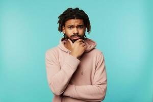 Puzzled young dark haired bearded guy with dreadlocks holding his chin with raised hand while looking pensively at camera, standing over blue background photo