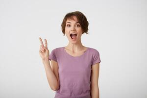 Funny young short-haired woman with wide open mout, makes peace sign with hand, expresses happiness, wears casual shirt poses indoor, stands over white wall. photo