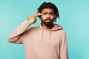 Sad young brown-eyed brunette bearded man with dark skin folding gun with raised hand and keeping it on his temple, wearing pink hoodie while posing over blue background photo