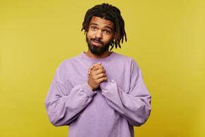 Indoor photo of cute young dark skinned brunette male with dreadlocks folding raised hands in thankful gesture and looking pleasantly at camera, posing over yellow background