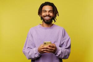 contento joven hermoso marrón peludo hombre con rastas sonriente alegremente a cámara mientras participación móvil teléfono, vestido en púrpura camisa de entrenamiento mientras siendo aislado terminado amarillo antecedentes foto