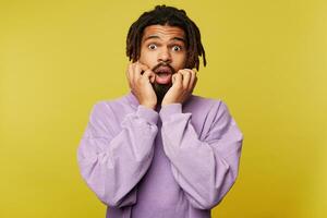 Portrait of shocked young attractive dark skinned brunette guy with beard holding raised hands on his cheeks while looking scaredly at camera, posing over yellow background photo