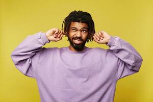 Displeased young dark skinned brunette bearded man with dreadlocks frowning his face while looking confusedly upwards and inserting forefingers into his ears, isolated over yellow background photo