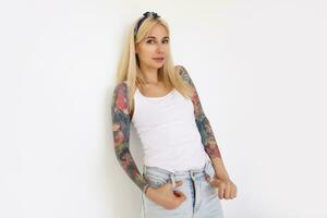 hermosa joven tatuado hembra con largo rubia pelo propensión su espalda en pared mientras posando terminado blanco antecedentes en camisa y vaqueros, sonriente ligeramente mientras mirando a cámara foto