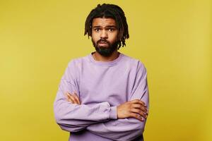 Indoor photo of young attractive dark skinned brunette guy with dreadlocks keeping his hands folded while looking sadly at camera, posing over yellow background