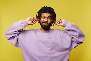 Dissatisfied young brown haired bearded male with dark skin covering his ears and grimacing face while trying to avoid annoying sounds, isolated over yellow background photo