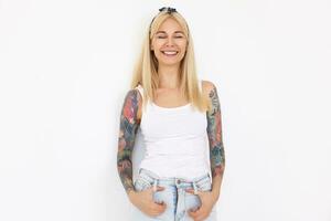 Good looking young happy long haired lady with tattoos wearing headband and casual clothes while posing over white background, keeping her eyes closed while smiling gladly photo