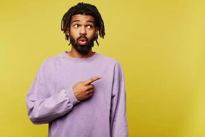 Confused young pretty brown haired bearded guy with dark skin showing surprisedly aside with raised index finger while standing over yellow background photo