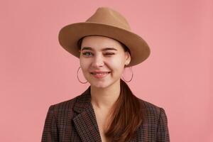 Close up of smiling attractive young woman, wearing in checkered jacket and brown hat, playful blinking. Looking at camera isolated over pink background. photo