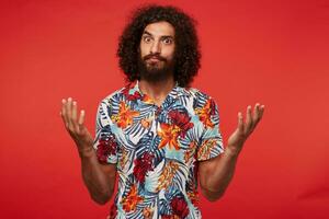 Studio photo of beautiful dark haired curly male with beard looking at camera with wide eyes opened and raising palms perplexedly, isolated against red background in multi-colored flowered shirt