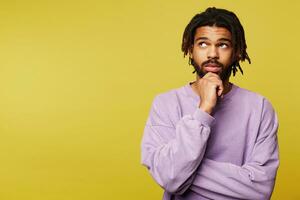 Pensive young brown eyed brunette bearded man with dark skin holding his chin with raised hand and looking thoughtfully upwards, isolated over yellow background photo