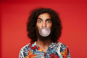 Amazed pretty brown-eyed bearded guy with dark curly hair making bubble with gum and looking surprisedly to camera, standing over red background in shirt with floral print photo