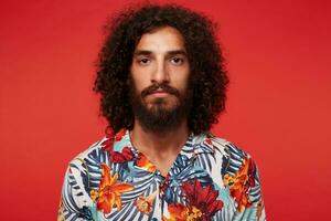 Portrait of handsome brown-eyed young bearded man with dark curly hair looking to camera with calm face and keeping his lips folded, isolated over red background in casual clothes photo