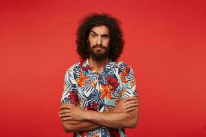 Bewildered handsome bearded brunette guy with curly hair keeping hands crossed on his chest while posing over red background, looking seriously to camera with raised eyebrow photo