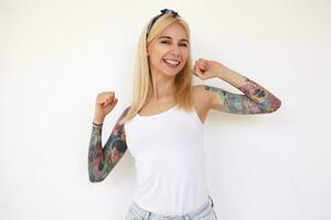 Studio photo of young cheerful tattooed blonde female with natural makeup smiling nicely to camera while stretching her hands, standing over white background