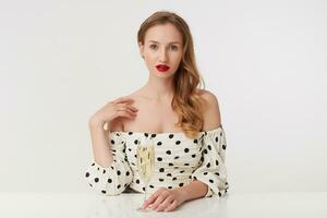 blue-eyed girl with long blond hair, with red lips in a polka-dot dress, sits at the table, puts her hand to her shoulder and holds a glass of champagne. Isolated over white white background. photo