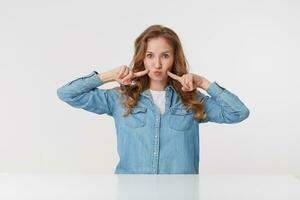 Photo of young cute woman with long blond wavy hair in denim shirt, touches his cheeks and blowing lips, looks funny. Isolated over white background.