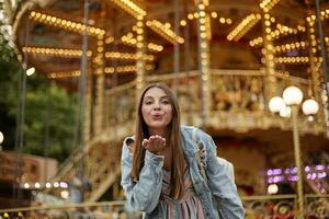 al aire libre foto de encantador largo peludo joven dama en de moda pantalones Saco levantamiento palma y soplo aire Beso a cámara, en pie terminado carrusel en parque de atracciones