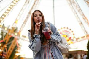 joven atractivo largo peludo hembra en pie terminado ferris rueda en parque de atracciones y Bebiendo limonada con paja, mirando a cámara y dando guiño foto