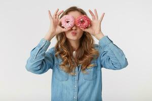 Portrait of young pretty woman with long blond wavy hair, wearing a denim shirt, looking through donuts and sending a kiss, isolated over white background. photo