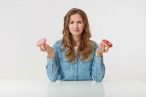 Displeased young woman with long blond wavy hair, sitting at the table and holds donuts in her hands. Frown, looking with disgust at camera isolated over white background. photo