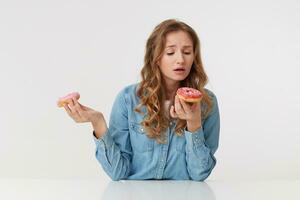 Photo of young beautiful woman with long blond wavy hair, wearing a denim shirt, holds donuts in her hands, gonna bite one of them. Isolated over white background.