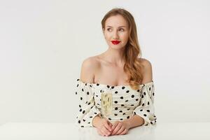 Photo of young beautiful blue-eyed girl with long blond hair, with red lips in a polka-dot dress, siting at the table with a glass of champagne, playfully looks away isolated over white background.