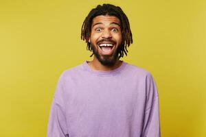 Joyful young attractive dark skinned brown haired man with dreadlocks looking surprisedly at camera with opened mouth while standing over yellow background photo