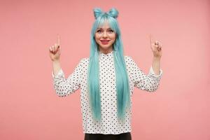 Cheerful young lovely blue haired female dressed in white shirt with black dots looking positively at camera and raising up forefingers, standing against pink background photo