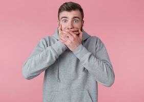 Photo of young handsome amazed red bearded man in gray hoodie, hears unbelievable news, looks surprised and covers mouth with palms, stands over pink background with wide open eyes.