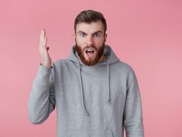 Portrait of young shocked red bearded man in gray hoodie, looks evil and displeased , with one raised hands, his favorite team lost. Stands over pink background and with wide open mouth. photo