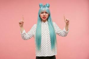 Displeased young blue haired lady frowning her face and looking at camera with pout, raising forefingers and pointing upwards, dressed in white shirt with black dots over pink background photo