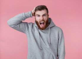 Portrait of young handsome shocked red bearded man in gray hoodie, looks evil and aggressive, stands over pink background and with wide open mouth. photo