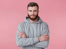 Portrait of young frowning handsome red bearded man in gray hoodie, stands with crossed arms, disapprovingly looks at camera, stands over pink backround. photo