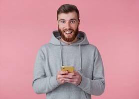 Young handsome positive red bearded man in gray hoodie, looks happy and broadly smiles, holding smartphone, stands over pink background. photo