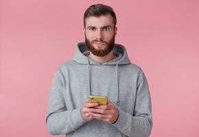retrato de joven atractivo rojo barbado hombre en gris sudadera, mira descontento, sosteniendo teléfono inteligente, mira a cámara y soportes terminado rosado antecedentes. foto