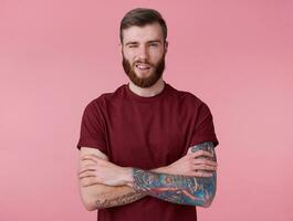 joven hermoso positivo rojo barbado chico en blanco camiseta, mira a cámara, guiño y en general sonrisas, soportes terminado rosado antecedentes y guiño. foto