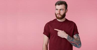 displeased young handsome red bearded man in red shirt, wants to draw your attention to copy space on the left side , pointing fingers and querulous looks at him, stands over pink background. photo