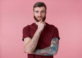 Portrait of young handsome thinking tattooed red bearded man in red t-shirt, touches the chin, stands over pink background looks at the camera. photo