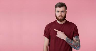 triste joven hermoso rojo barbado hombre en rojo camisa, quiere a dibujar tu atención, señalando dedos a el izquierda a el Copiar espacio, soportes terminado rosado antecedentes con labios abajo. foto