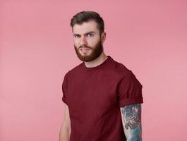 Photo of young handsome frowning misunderstanding red bearded man in blank t-shirt, stands over pink background looks at the camera with disgust.