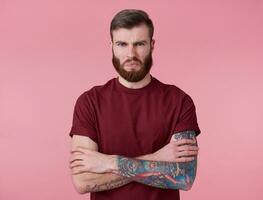 Photo of young handsome tattooed disgusted red bearded man in blank t-shirt, stands with crossed arms over pink background, frowning and looks at the camera.