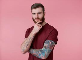 Photo of young attractive thinking tattooed red bearded man in red t-shirt, stands over pink background, looks at the camera and touches the chin.