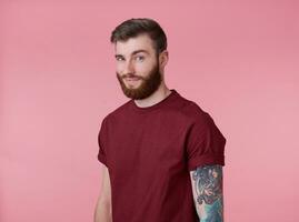 Portrait of young handsome happy red bearded tattooed man in red t-shirt, looks cheerful and broadly smiles, looks away, stands over pink background. photo