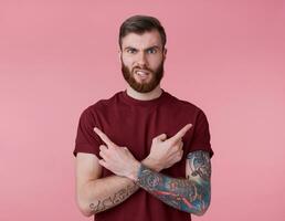 Portrait of young handsome frowning misunderstanding red bearded man in red t-shirt, stands over pink background looks at the camera with disgust and points in different directions. photo