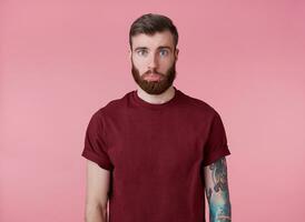 retrato de triste joven hermoso rojo barbado tatuado hombre en rojo camiseta, ofendido mirando a el cámara con un bajado labio, soportes terminado rosado antecedentes. foto