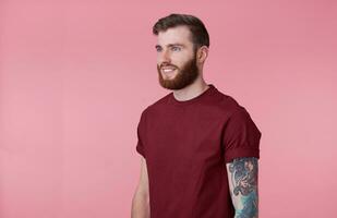 Photo of young handsome cheerful red bearded tattooed man in red t-shirt, looks happy and broadly smiles, looks away, stands over pink background.