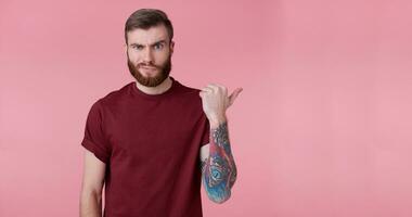 young handsome tattooed misunderstanding red bearded man in blank t-shirt, stands over pink background, looks at the camera and wants to draw you attention and points to copy space on the right side. photo